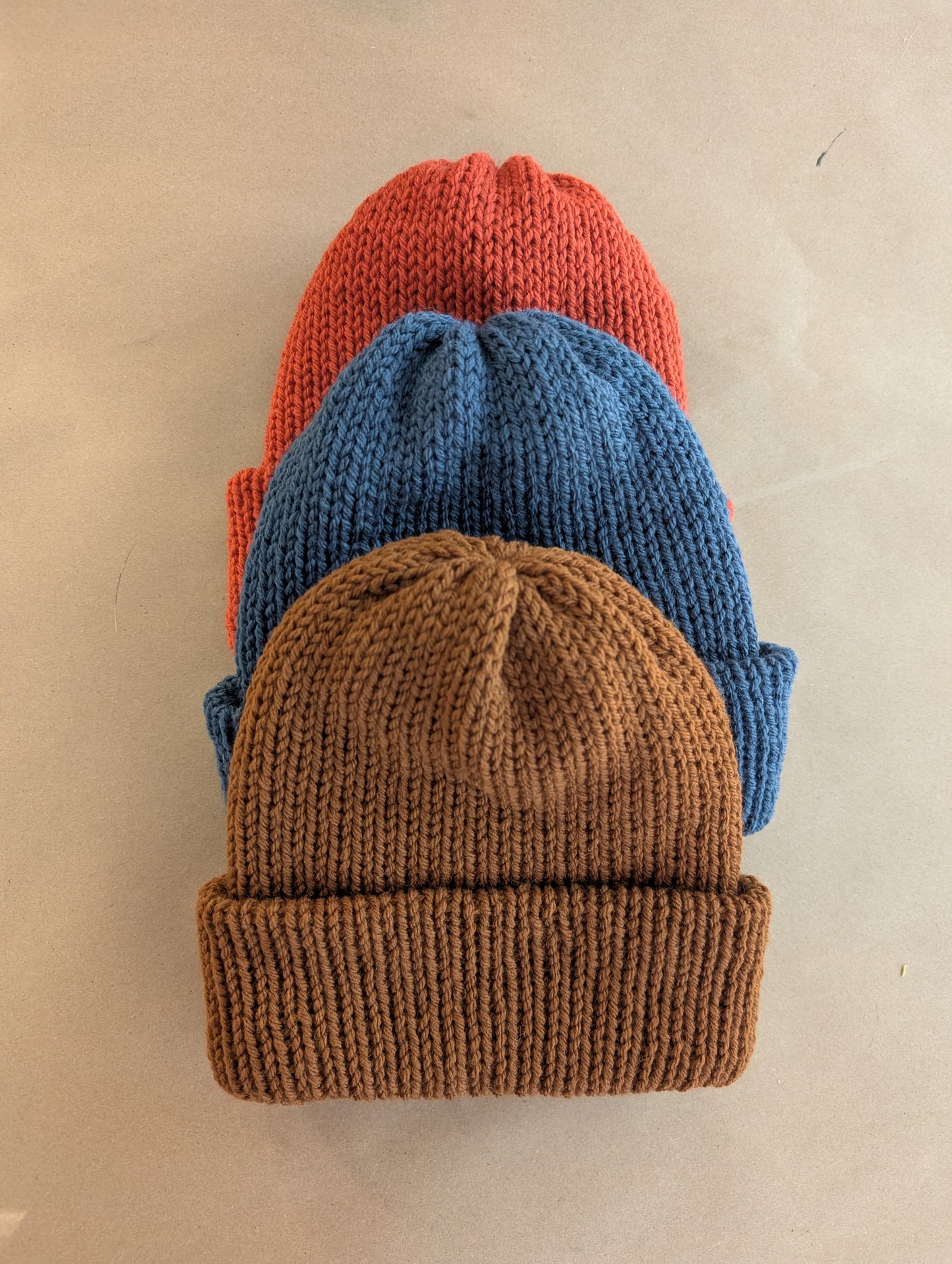 Three knit beanie hats in orange, blue, and brown stacked on a beige background.