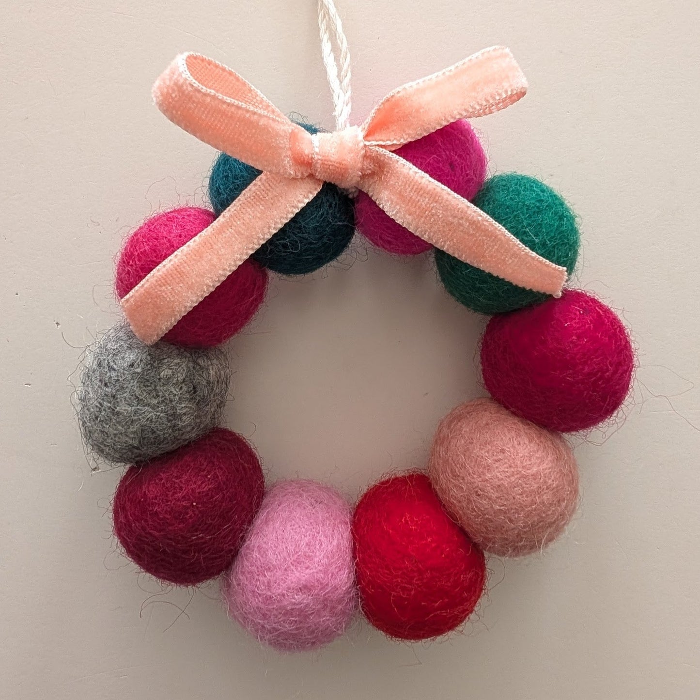 Decorative wreath ornament with pink, red, green, and grey wool balls, and a pink bow on a white background.