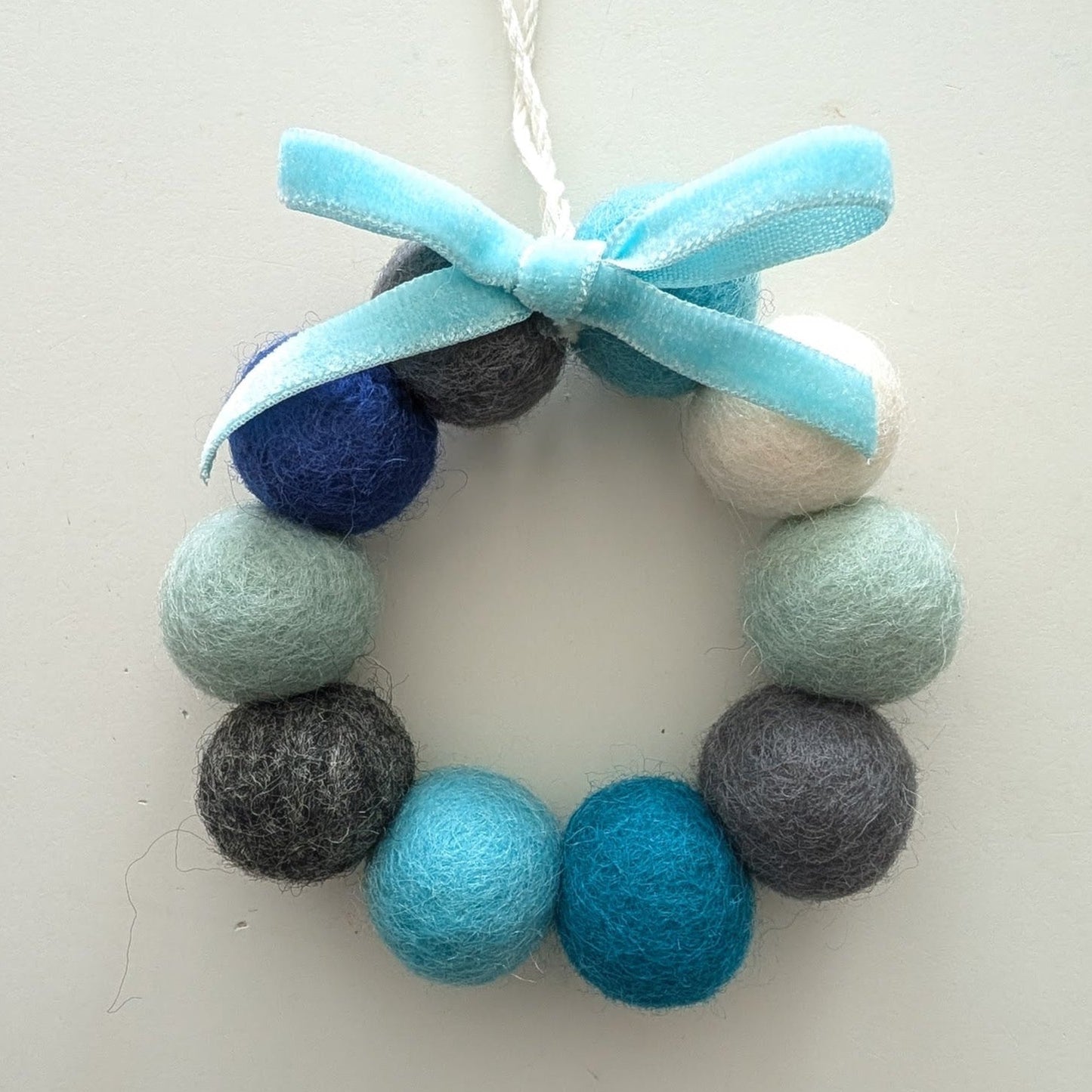 Decorative wreath with blue, grey, and white felt balls and a blue bow on a white background.