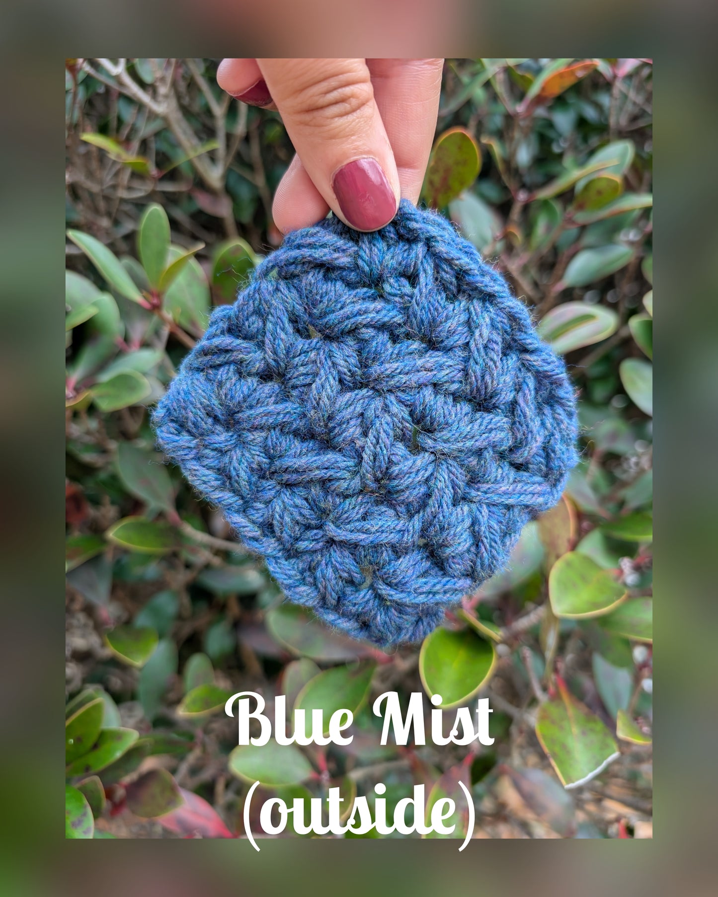 Blue crocheted item held by a hand with green foliage in the background