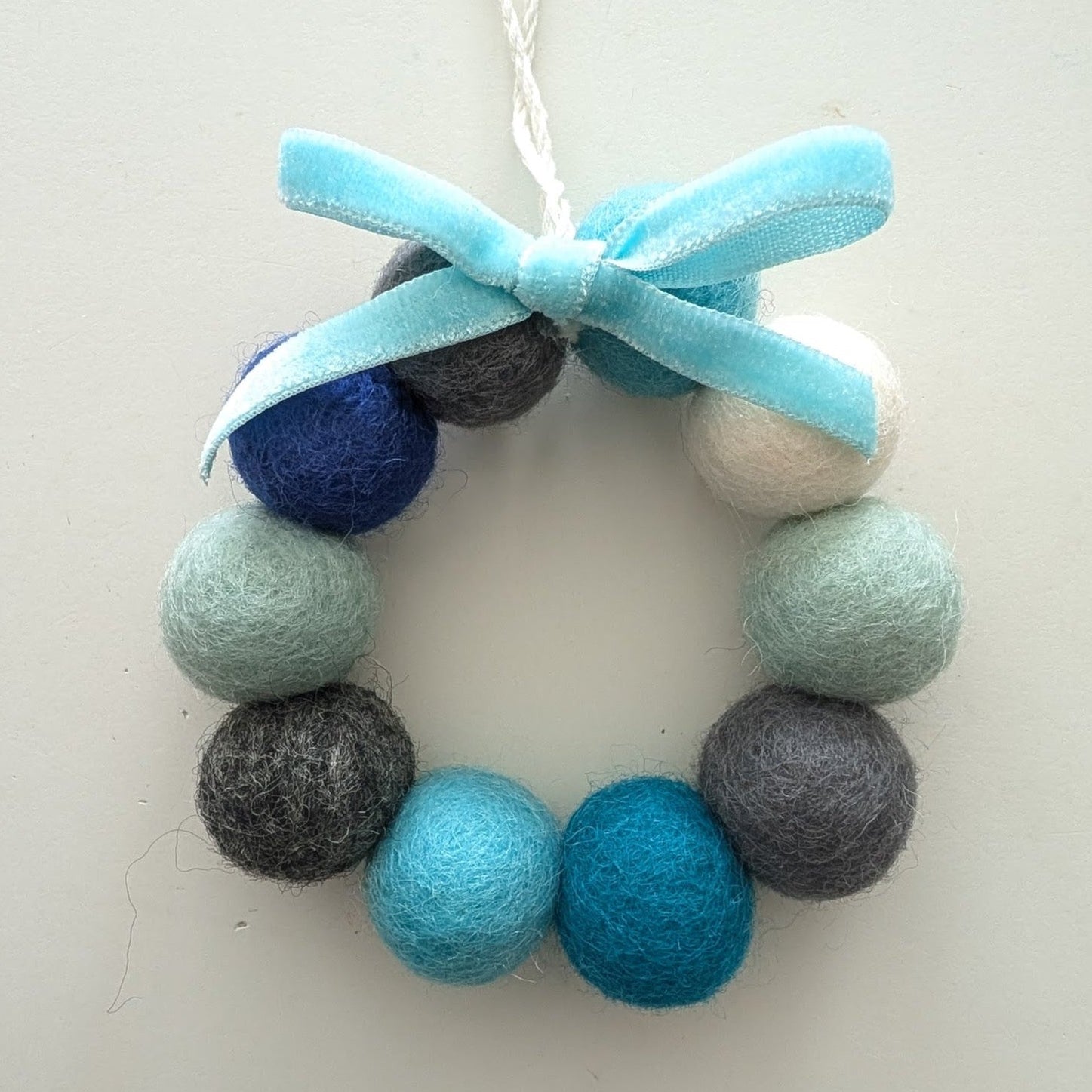 Decorative wreath with blue, grey, and white felt balls and a blue bow on a white background.