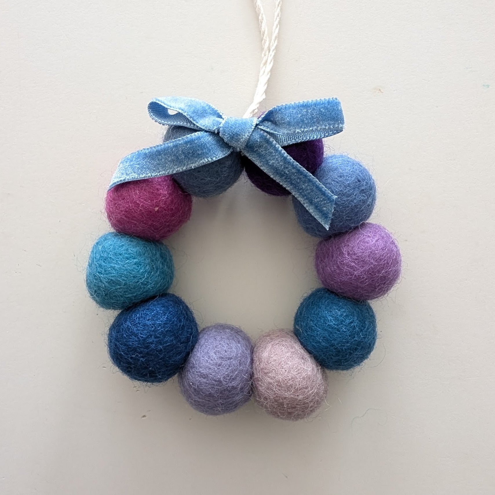 Decorative wreath ornament with blue and purple felt balls and a bow on a white background.